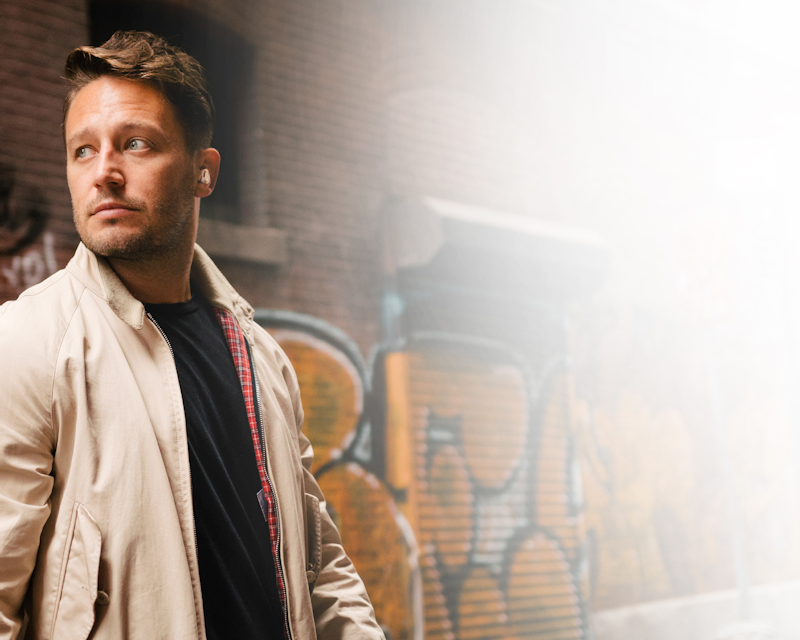 Man standing in front of a graffiti covered brick wall wearing a beige jacket and Klipsch T5 II True Wireless ANC Earphones desktop