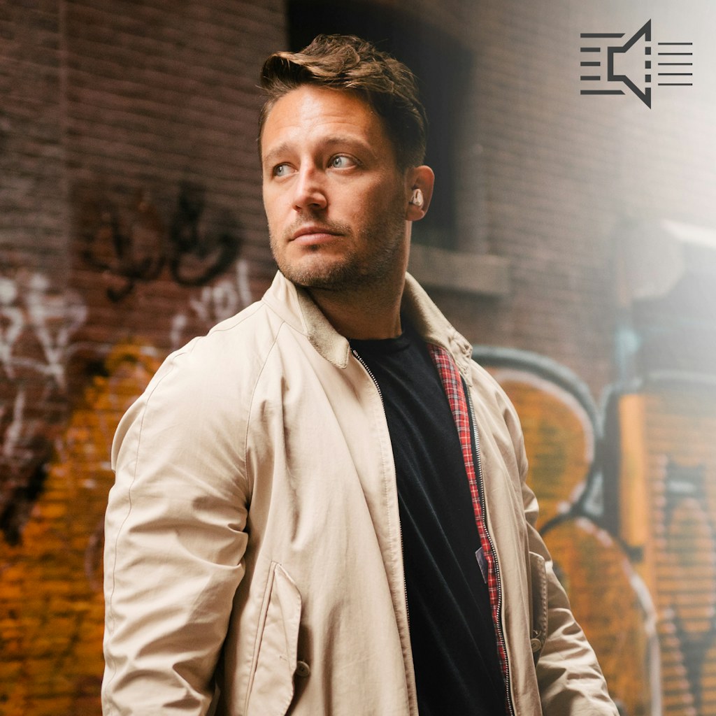 Man standing in front of a graffiti covered brick wall wearing a beige jacket and Klipsch T5 II True Wireless ANC Earphones mobile